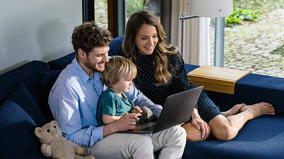[Translate to English:] A man, a woman and a child are sitting on a sofa in front of a laptop.