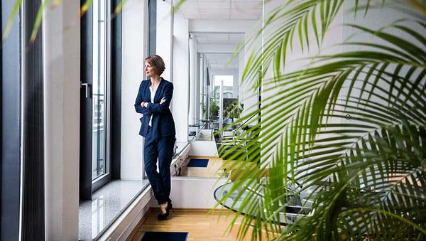 Woman in dark blue suit leaning with folded arms at the window in a modern office with plants